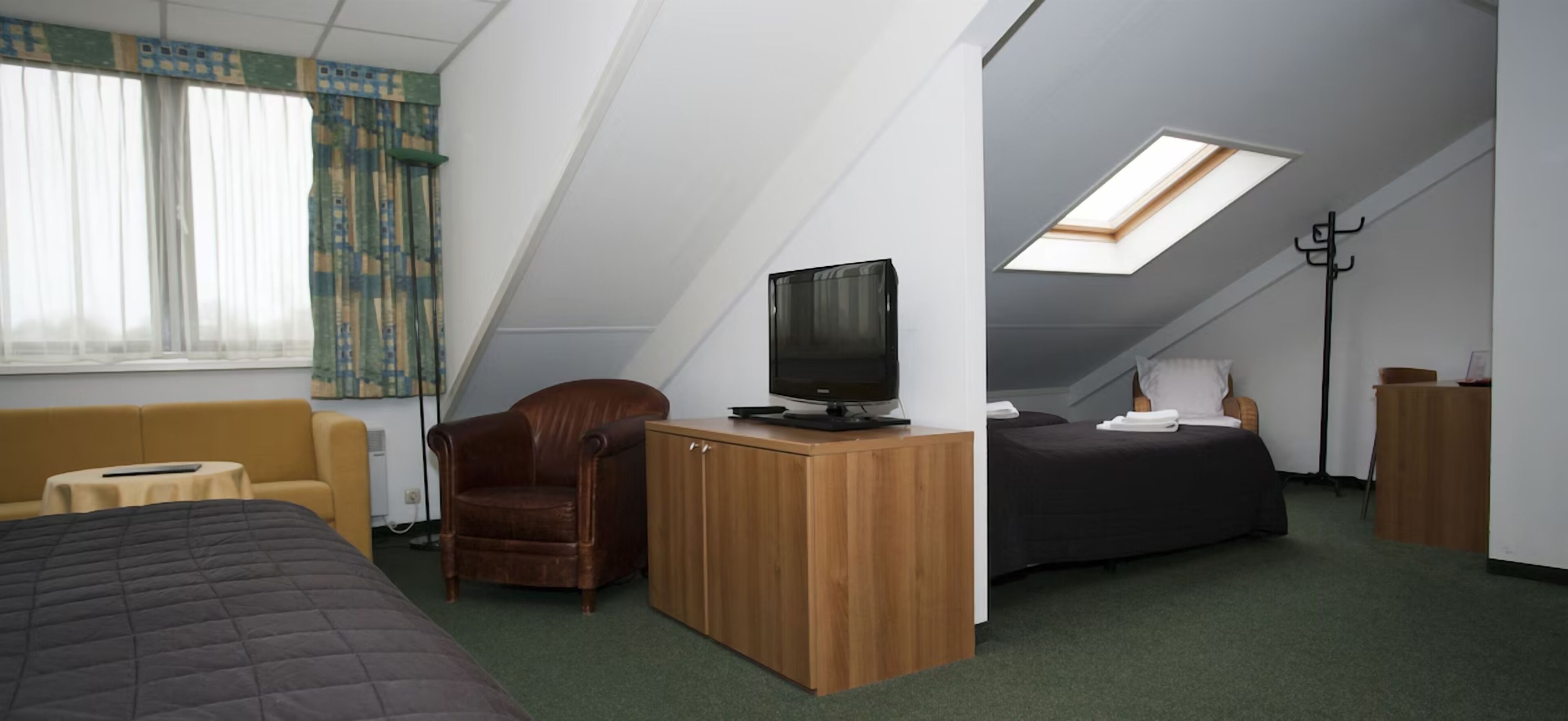 A compact hotel room with a bed, a sofa, a desk with a TV, a chair, a bedside table, a lamp, and a skylight.