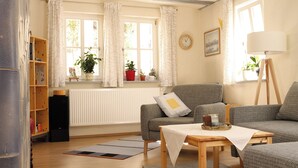 A cozy living room with a sofa, a small table, a bookshelf, a radiator, and a window with curtains. - Historic wine farmhouse in Main/Wine Franconia at the foot of the Spessart (Triefenstein)