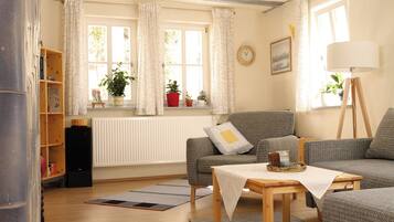Ein gemütliches Wohnzimmer mit einer Couch, einem kleinen Tisch, einem Bücherregal, einer Heizung und einem Fenster mit Vorhängen.