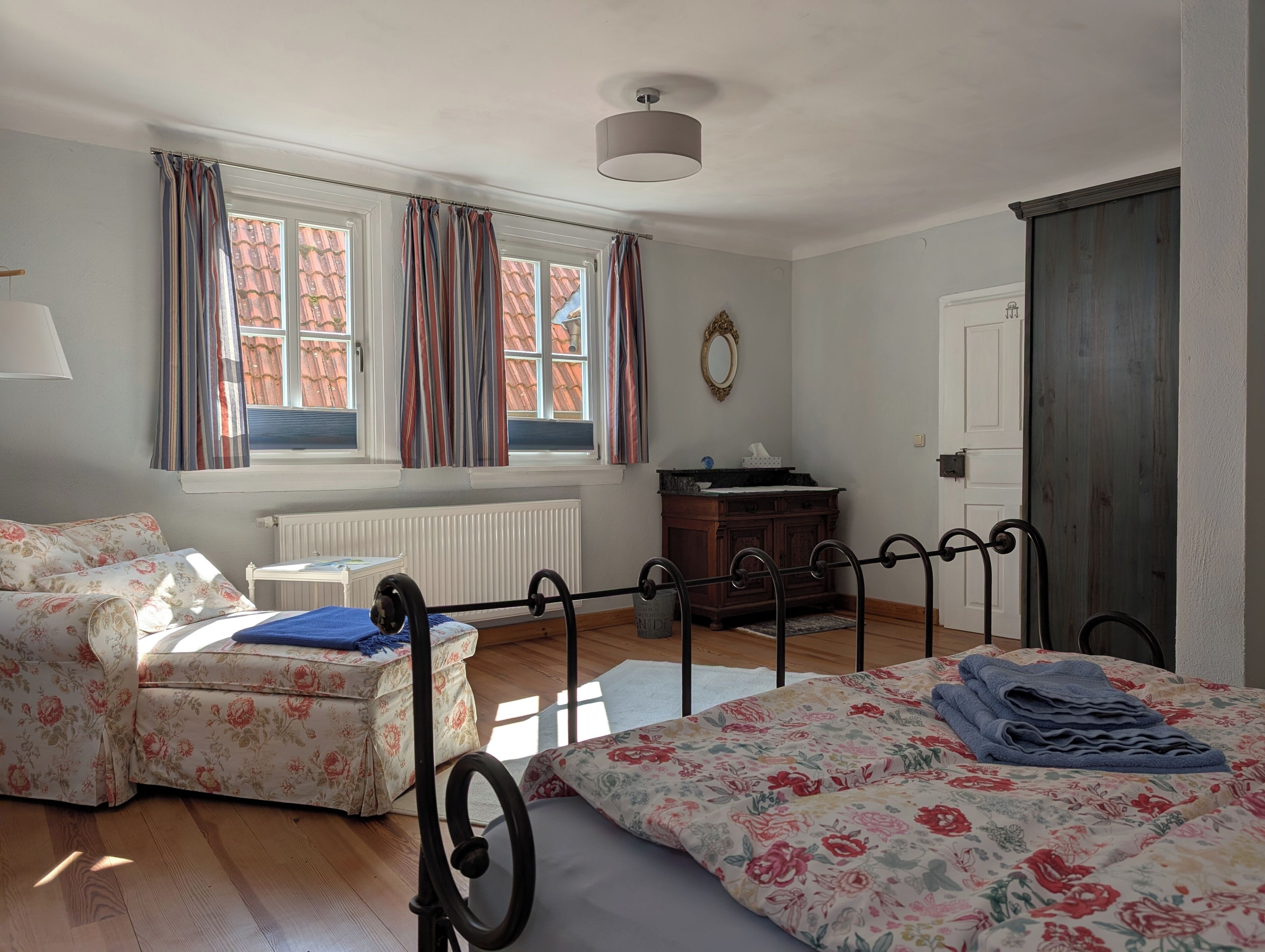 Ein Schlafzimmer mit Bett, Sofa, Schminktisch und Heizkörper, sowie einem Fenster mit Vorhängen.
