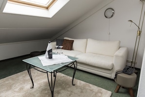 Een kamer met een tafel met glazen blad, een witte bank, een klein rond tafeltje met een telefoon en een vloerlamp.