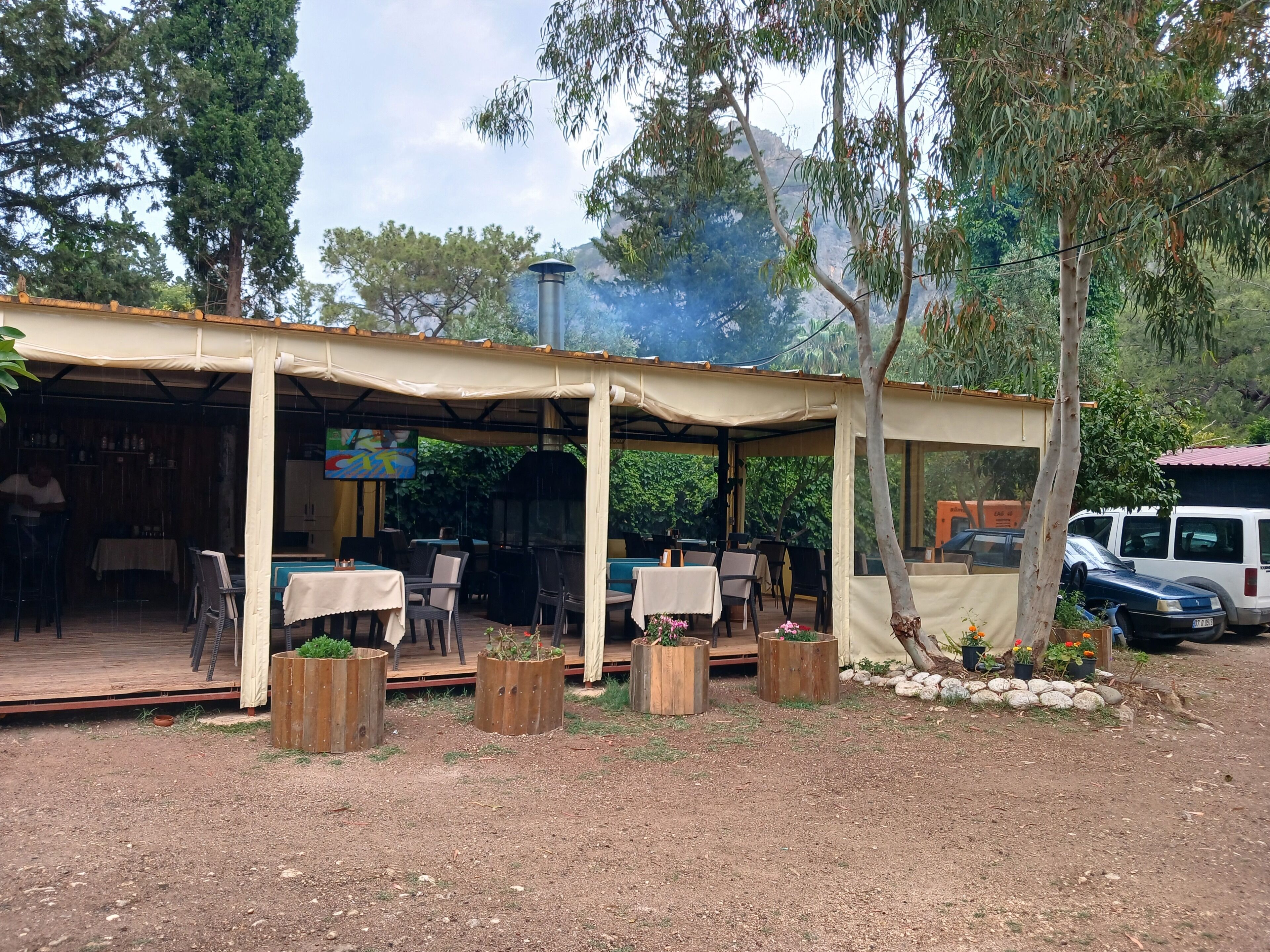 Un área de comedor al aire libre con mesas y sillas, rodeada de árboles y una estructura cubierta.