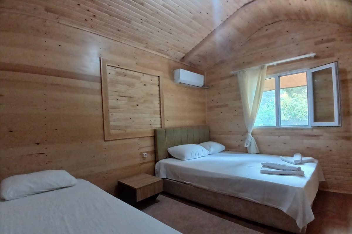 A wooden cabin room with two beds, a window with curtains, and a wall-mounted air conditioning unit.