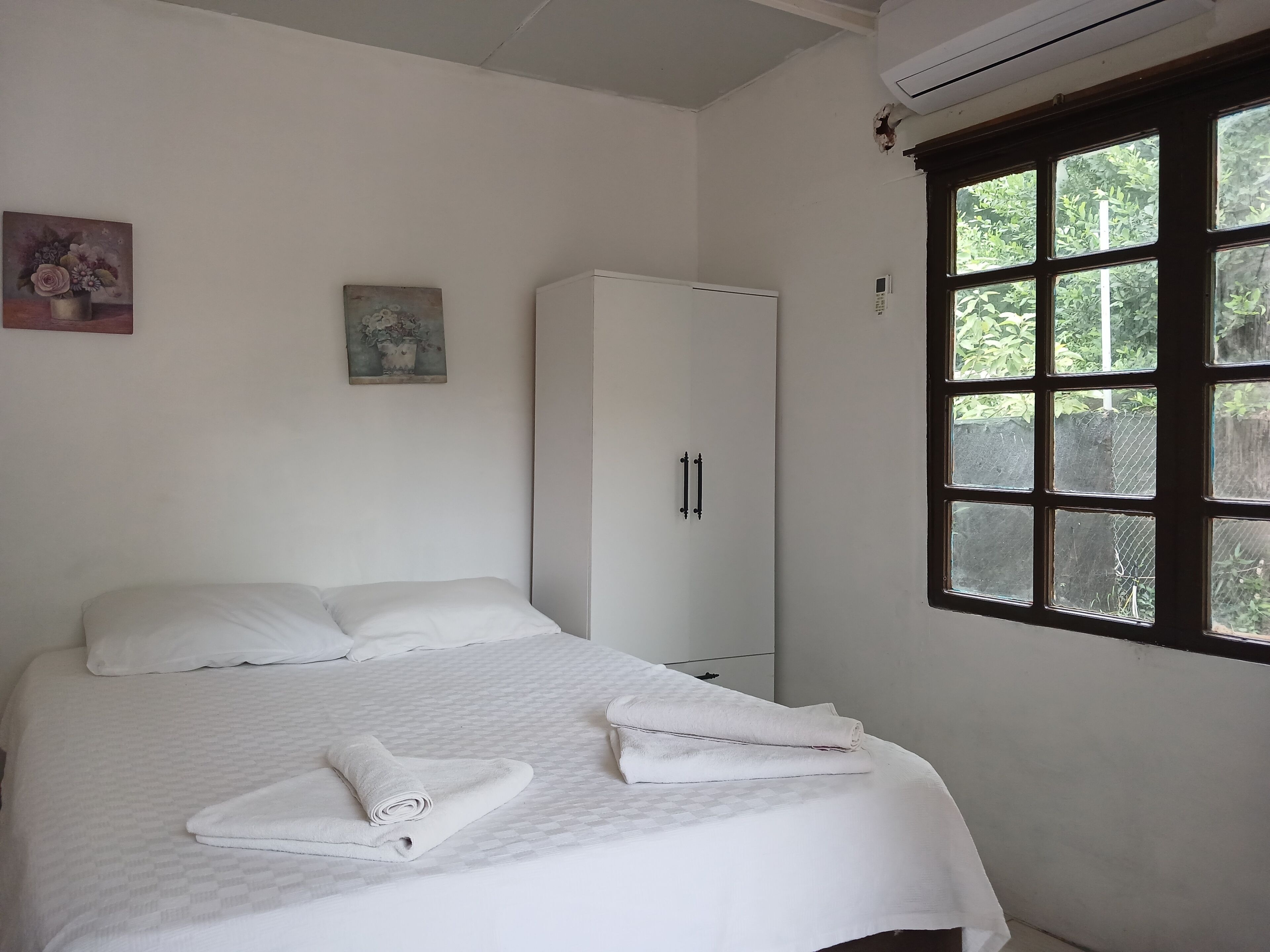 A neatly made bed with white linens, a white wardrobe, and a window with a view of greenery.