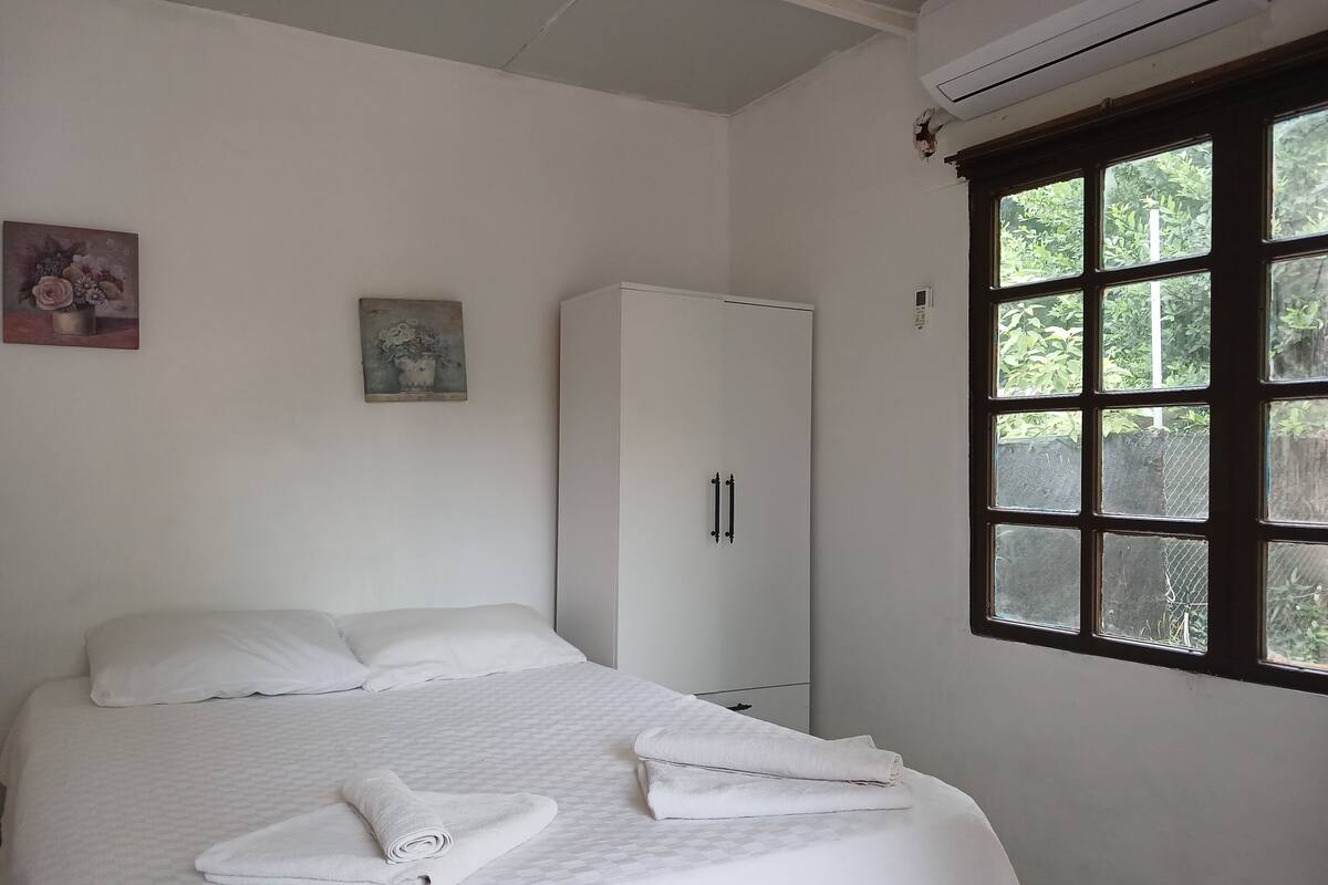 A neatly made bed with white linens, a white wardrobe, and a window with a view of greenery.