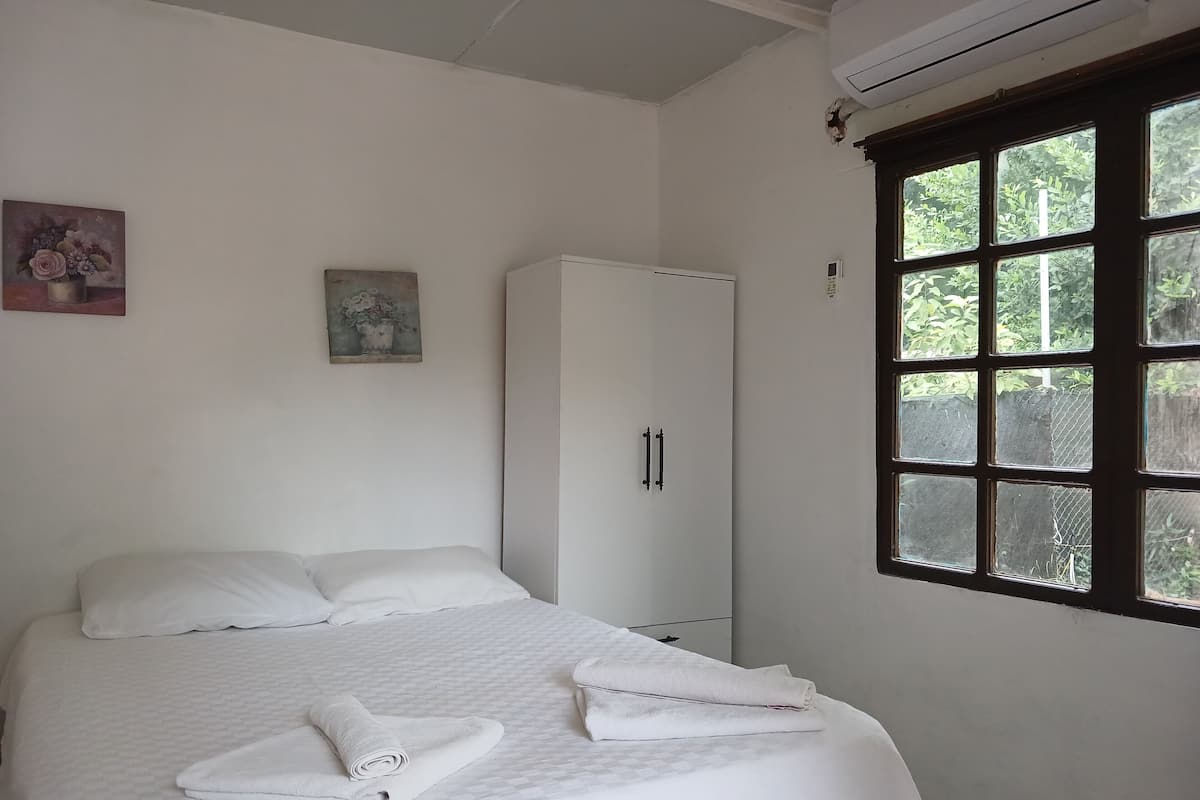 A neatly made bed with white linens, a white wardrobe, and a window with a view of greenery.