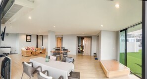 Ampla sala de estar com mesa de jantar, móveis de vime e vista para um gramado verde por grandes janelas.