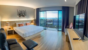 Ein modernes Hotelzimmer mit einem großen Bett, einem Schreibtisch, einem Sessel, einer Couch und einem Balkon mit Stadtblick.