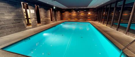 An indoor swimming pool with a tiled floor, a brick wall with a clock, and a glass partition on one side.