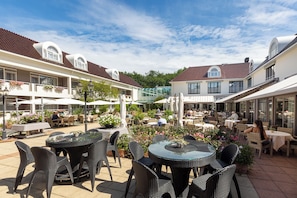 An outdoor patio area with round tables and wicker chairs, surrounded by buildings with red-tiled roofs and large windows. - Standard plus double room (Zeist)