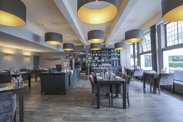 A modern restaurant interior with high ceilings, pendant lighting, a central bar area, and a view through large windows.