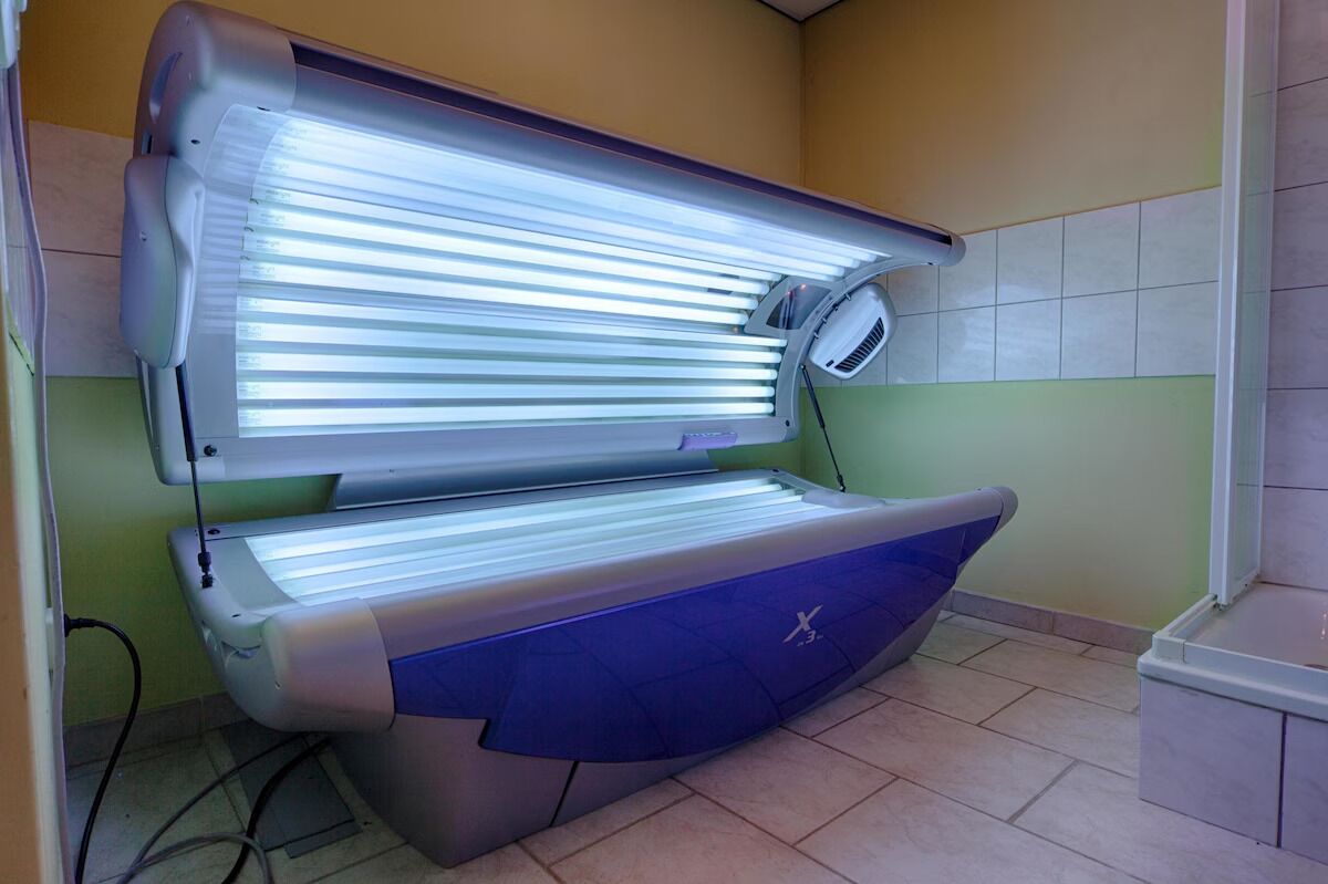 A tanning bed with a control panel, a footrest, and a wall-mounted fan.