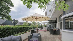 Een terras buiten met rieten meubels, een glazen tafel en een grote parasol.