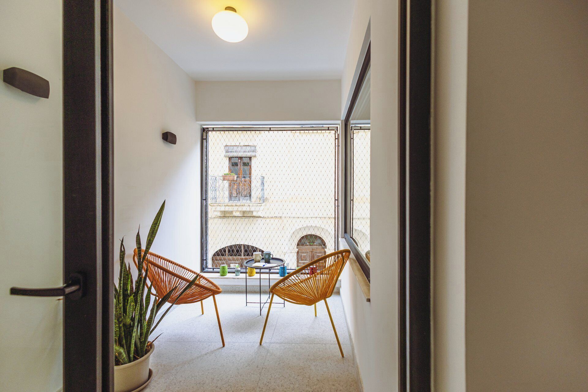A narrow hallway leading to a balcony with two wicker chairs and a potted plant.