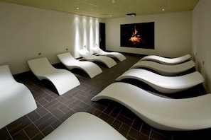 Une pièce comprenant plusieurs fauteuils de massage blancs disposés en rangées, un grand écran mural diffusant une image de feu, et un sol carrelé.