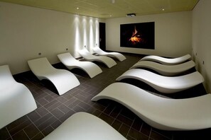 A room with multiple white massage chairs arranged in rows, a large wall-mounted screen displaying a fire, and a tiled floor.