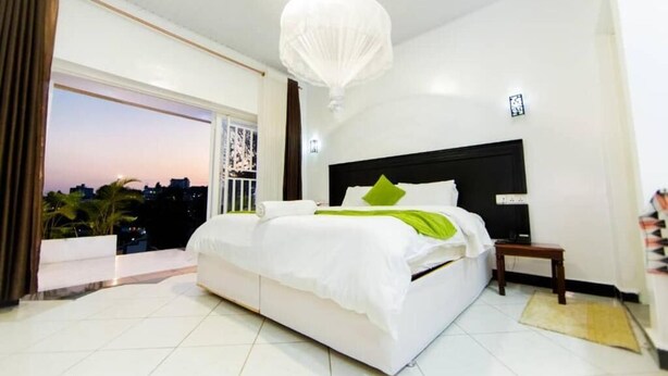 A bedroom with a large bed, a nightstand, a white ceiling light, and a view of the city at sunset. - Inzozi Africa House B&B (Kigali)