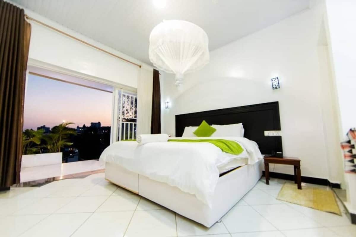 A bedroom with a large bed, a nightstand, a white ceiling light, and a view of the city at sunset.