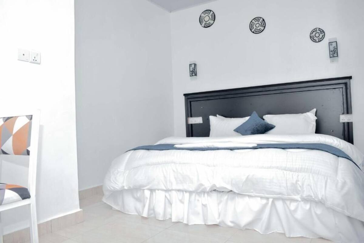 A neatly made bed with white bedding and a dark headboard, a white chair with a geometric cushion, and a wall with three decorative plates.