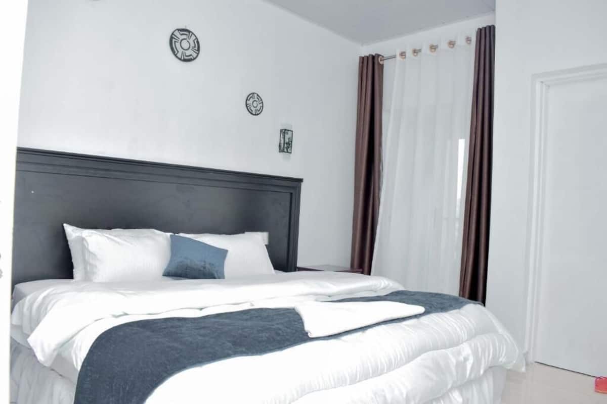 A neatly made bed with a dark headboard, white bedding, and a blue accent pillow. There are two wall decorations and a window with curtains.