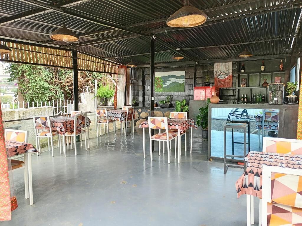 Un espacio de estar al aire libre cubierto con mesas y sillas, una zona de bar con estantes llenos de botellas y un mural en la pared.