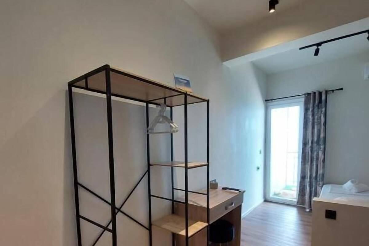 A room with a wooden floor, a black metal shelving unit, a desk with a chair, and a bed with a headboard.