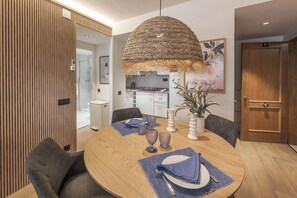 A dining area with a round table, chairs, a hanging light fixture, and a kitchen in the background.