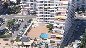 Un hotel de altura con varios balcones, un estacionamiento y una zona de playa con sombrillas.