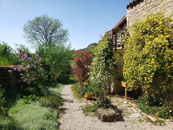 Un étroit sentier de gravier mène à un bâtiment en pierre doté d’un balcon en bois, le tout étant entouré d’une végétation luxuriante et de plantes en fleurs.