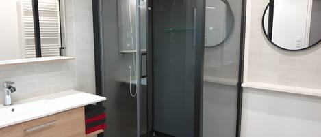 Een moderne badkamer met een douchecabine van glas, een witte wastafel met chromen kraan en een ronde spiegel aan de wand.