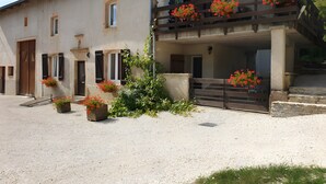 A two-story building with a gravel driveway, a wooden gate, and potted plants with red flowers. - Gîte de la Verdurette: Tranquil Stay in the Countryside (Pettonville)