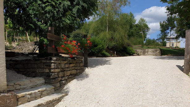 A gravel pathway, stone wall, wooden cross, and red flowers. - Gîte de la Verdurette: Tranquil Stay in the Countryside (Pettonville)