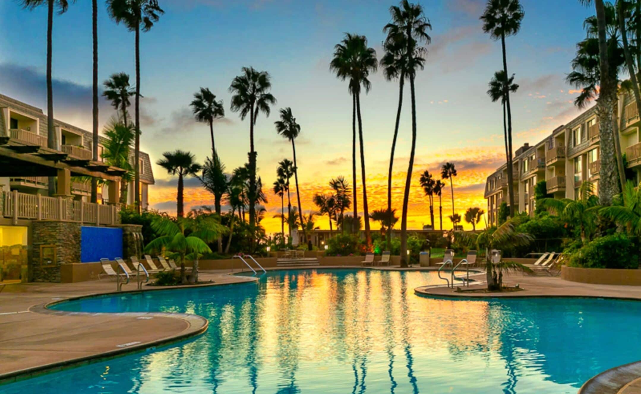 Una piscina con sillones de descanso, palmeras y edificios de hotel al atardecer.