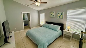 Une chambre à coucher comprenant un lit, un ventilateur de plafond, une télévision, une table de chevet, une lampe et une fenêtre avec des stores.