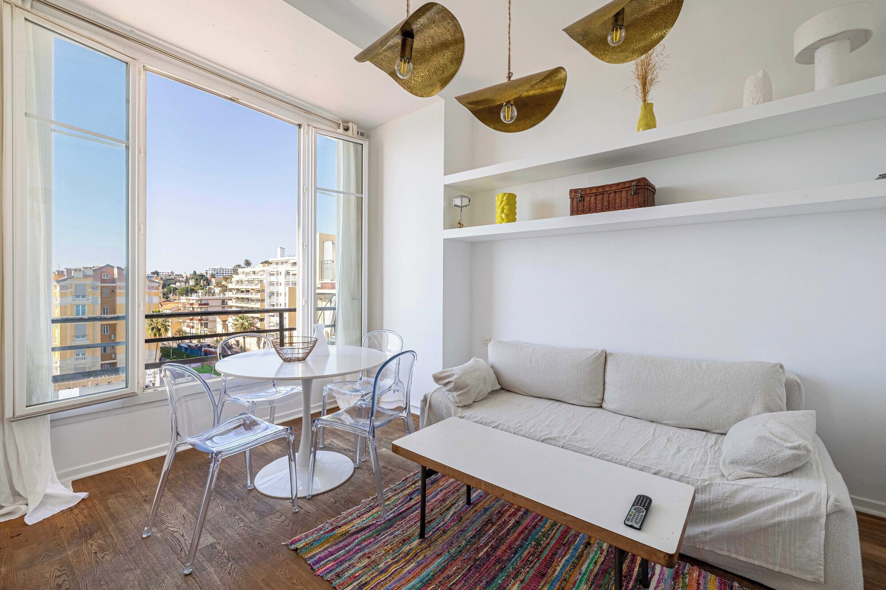 Una sala de estar moderna con sofá, una mesa de centro, una mesa de comedor con sillas y un gran ventanal con vistas a edificios.