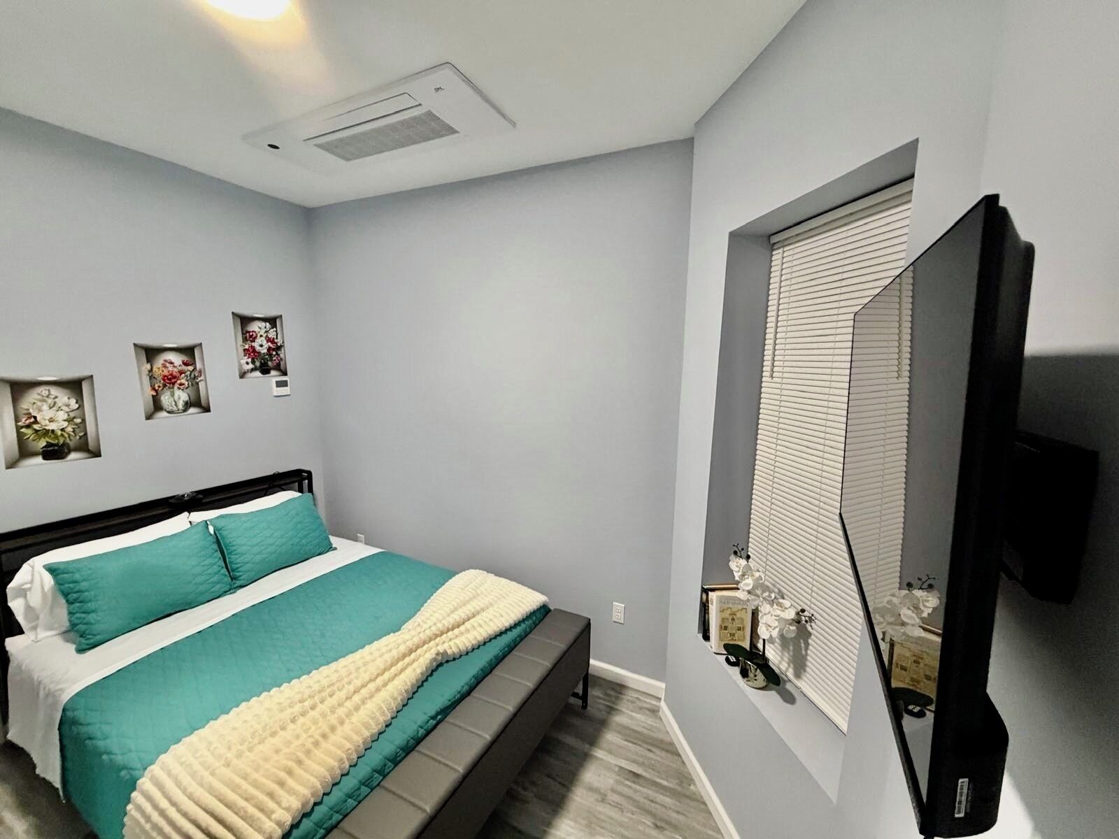 Una camera da letto con un letto, una TV a parete, una finestra con persiane e una bocchetta di aerazione al soffitto.
