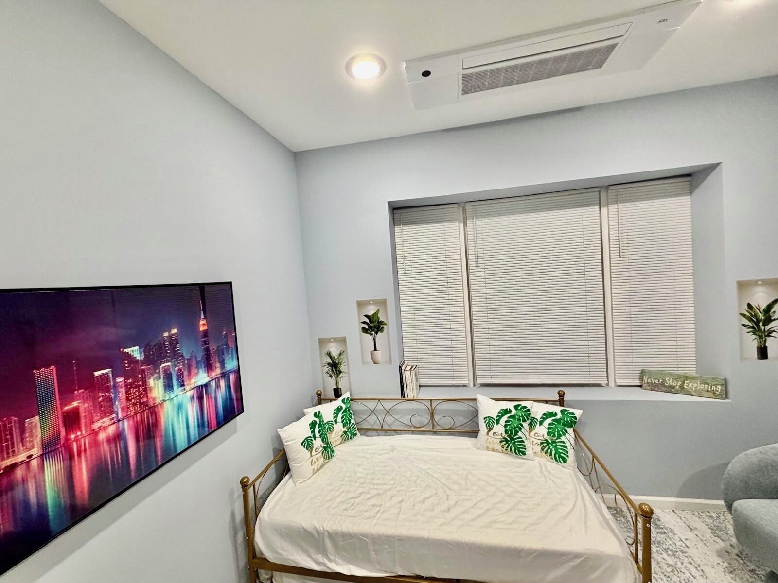 A bedroom with a bed, a cityscape painting, a window with blinds, and a motivational sign.