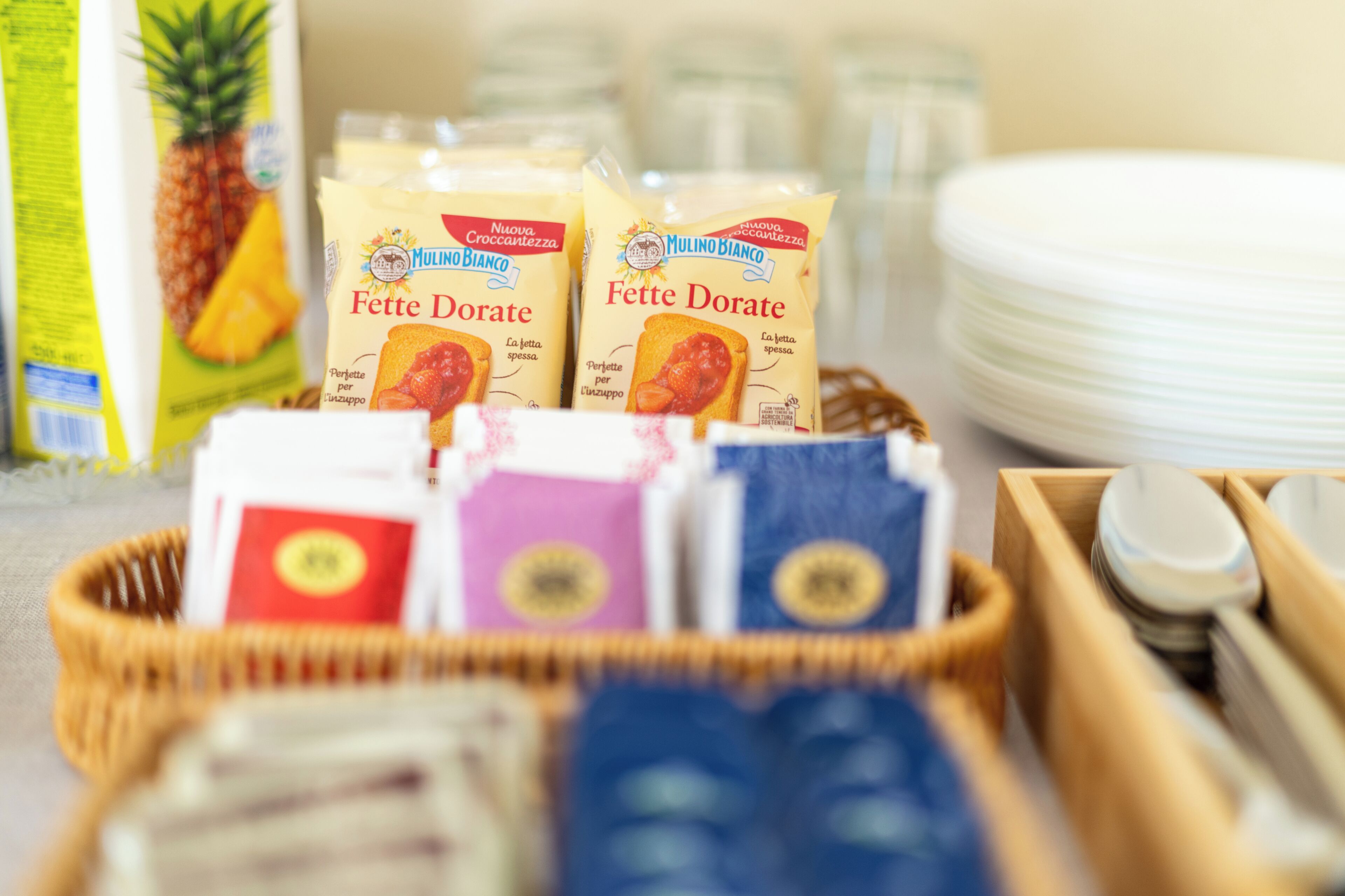 A breakfast setting with a wicker basket containing two packages of Fette Dorate, a carton of milk, and a pineapple.