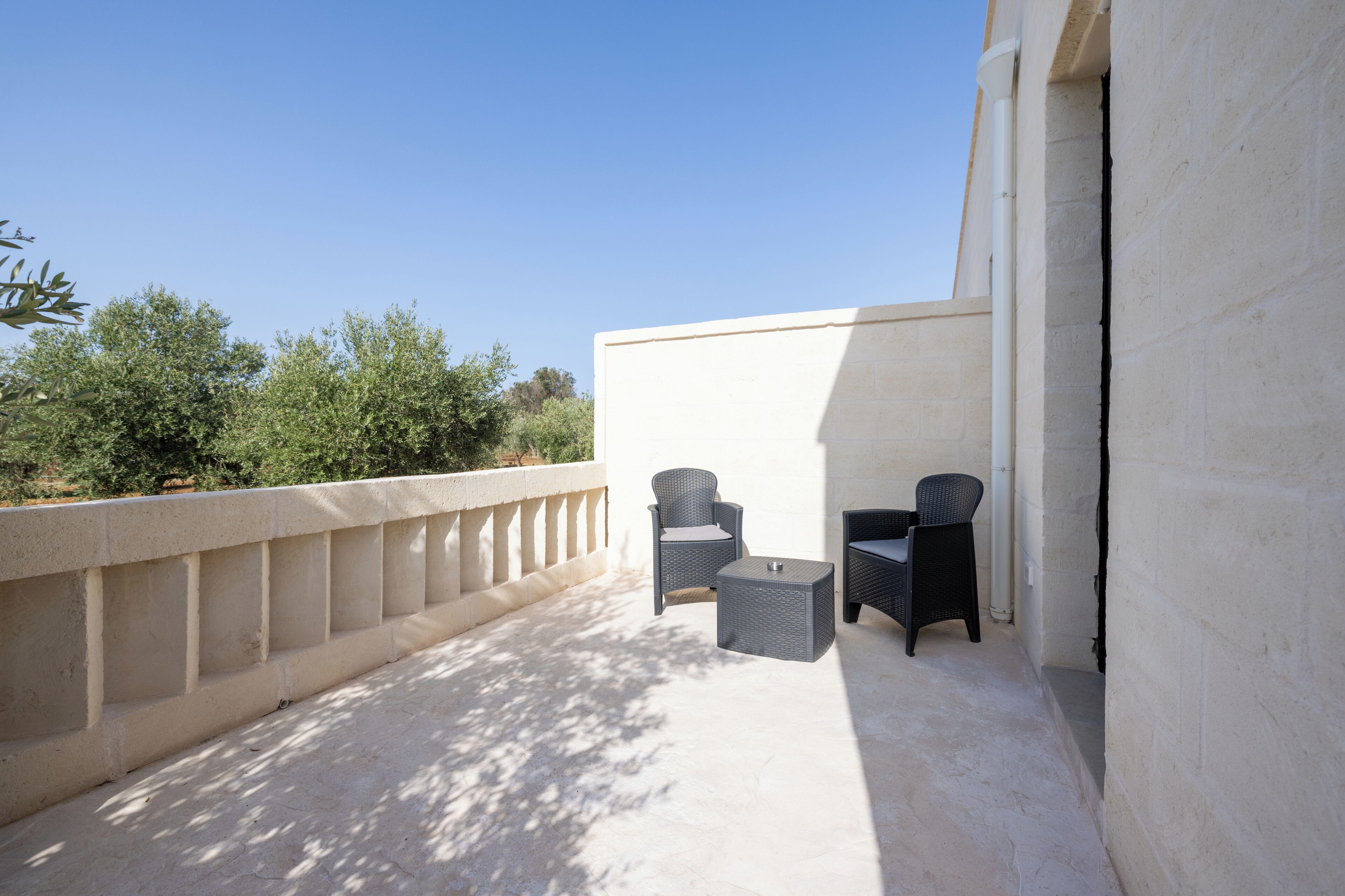 Un área exterior con asientos, dos sillas de mimbre y una pequeña mesa, suelo de hormigón y pared blanca.