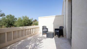 Un área exterior con asientos, dos sillas de mimbre y una pequeña mesa, suelo de hormigón y pared blanca.