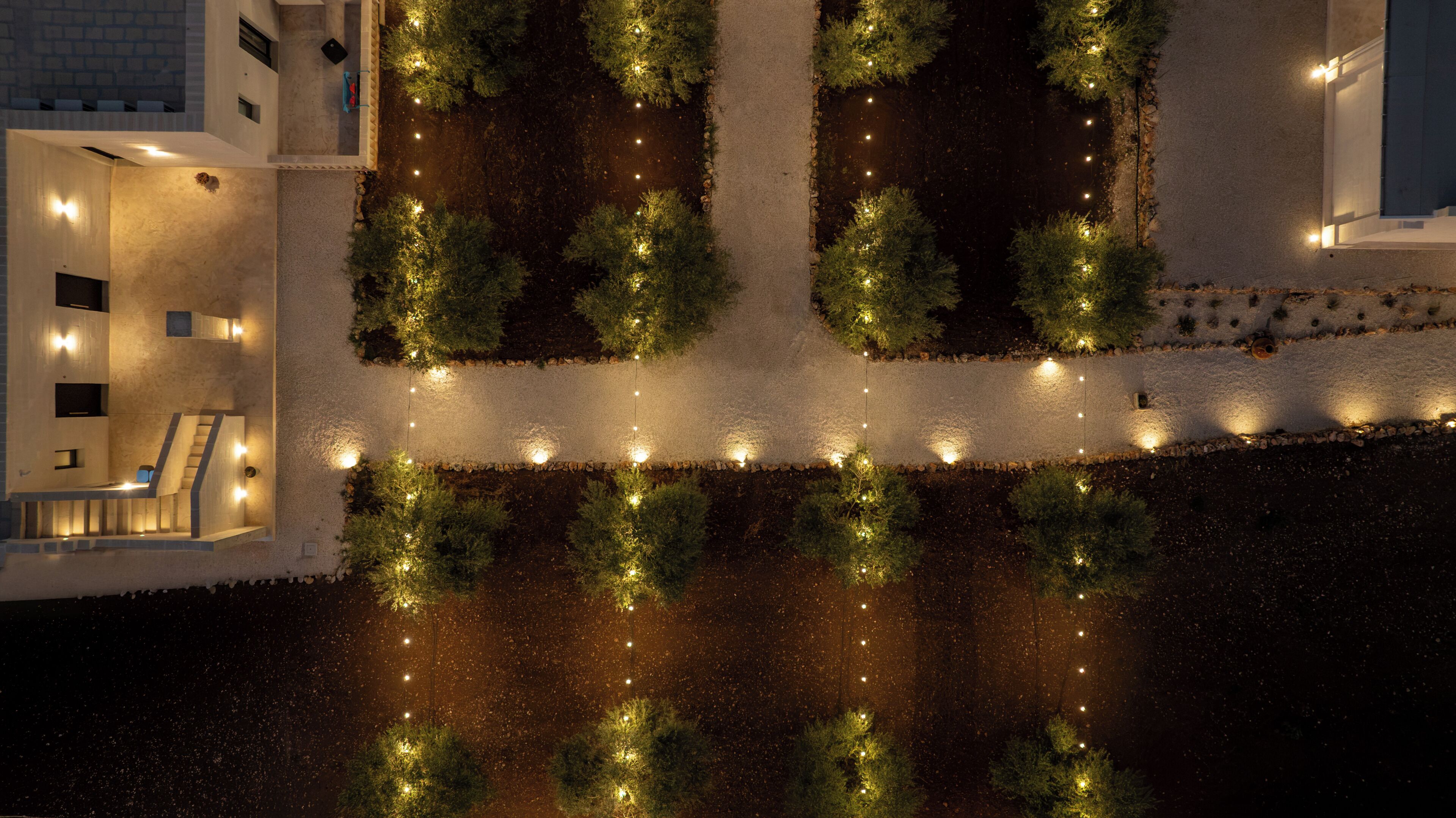 Vista cenital de un jardín ajardinado con un sendero, bordeado por árboles regularmente espaciados e iluminado por luces en el suelo.