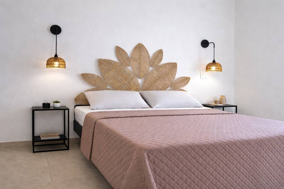 Un lit avec une tête de lit ornée d’un motif de feuilles, deux tables de chevet avec des lampes, une plante sur l’une des tables de chevet et une couverture de lit rose matelassée.