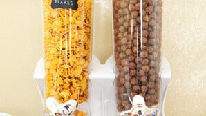 Due distributori di vetro trasparente su un supporto bianco, uno con l'etichetta 'CORN FLAKES' e l'altro 'CORN FLOUR', con una parete beige sullo sfondo.