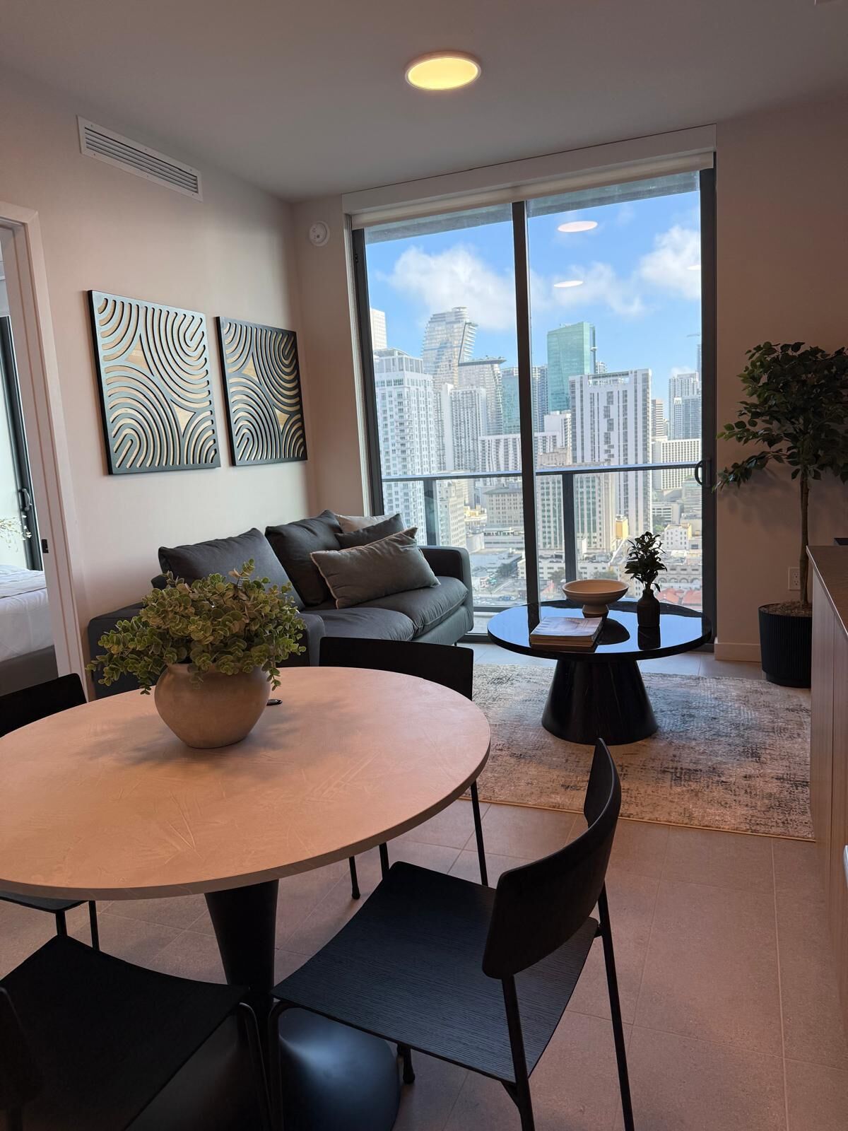 A modern living room with a round table, two black chairs, a grey sofa, and a large window offering a city view.