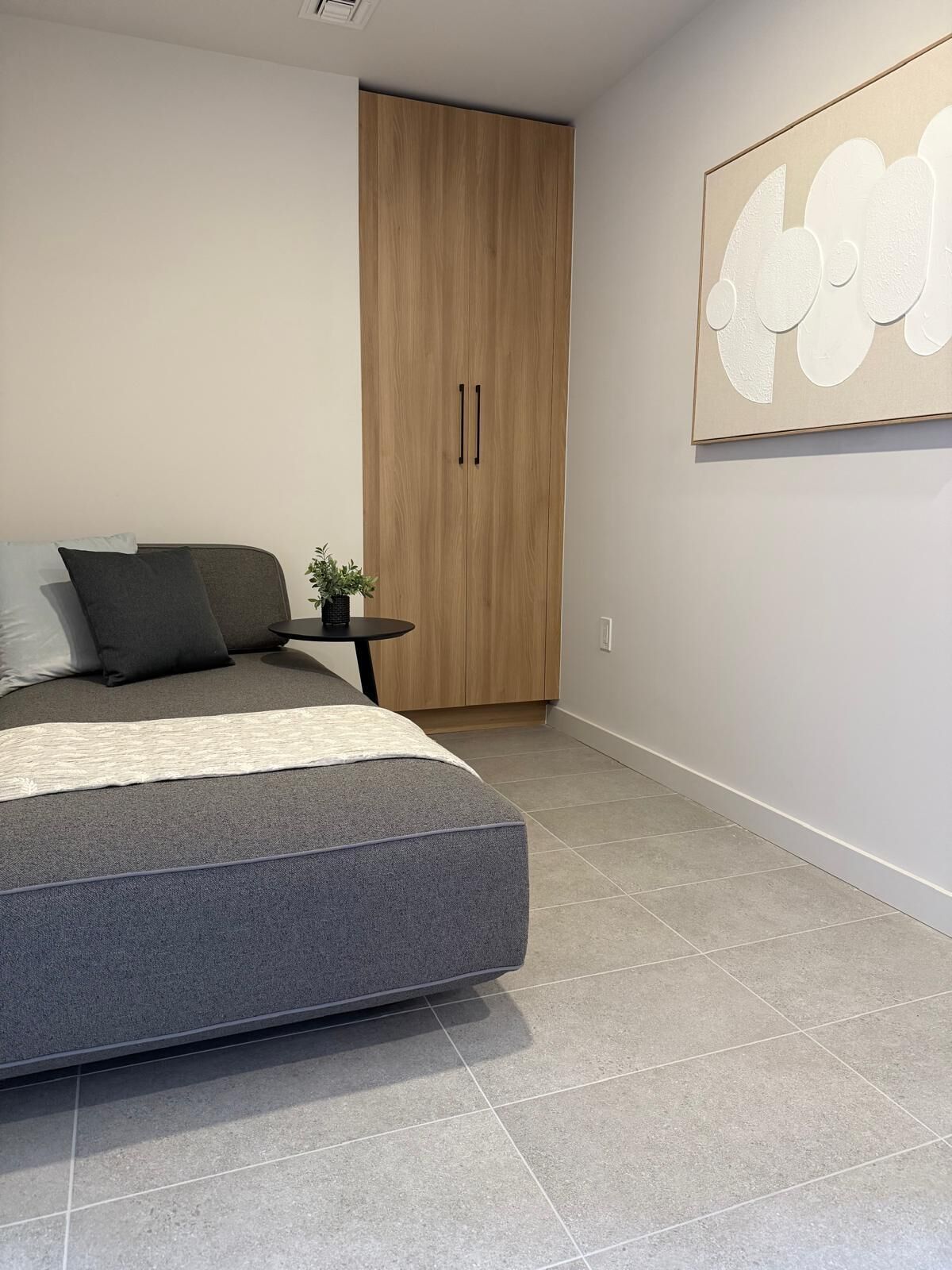 Un dormitorio moderno con un colchón gris, un armario de madera, una mesita con una planta y una obra de arte en la pared.
