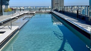 En pool på taket med kristallklart blått vatten, omgiven av moderna byggnader och en klar himmel.