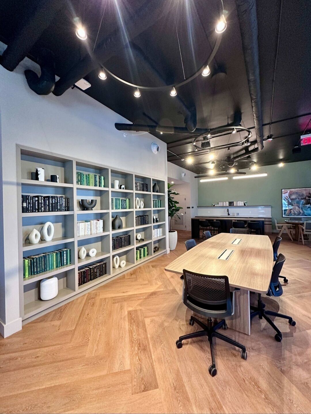 Ein moderner Büroraum mit einem großen Holztisch, schwarzen Bürostühlen und einem weißen Bücherregal mit Büchern und Dekorationsobjekten.