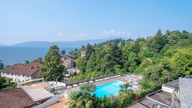 Ein Poolbereich mit Liegestühlen, Blick auf das Meer und Gebäude, die von Bäumen umgeben sind.