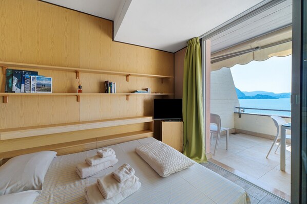A hotel room with a bed, a TV, a wooden shelf with books, and a balcony with a view.