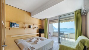Ein Hotelzimmer mit Bett, einem grünen Sessel, einem Fernseher, einem hölzernen Bücherregal und Meerblick.